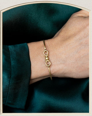 A delicate gold bangle bracelet with a central bead and ring motif, shown worn on a person's wrist against a dark teal satin fabric background.