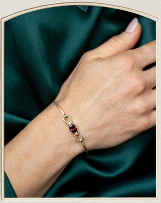 A close-up of a person's wrist wearing a delicate gold bracelet with a central dark red oval gemstone, set against a dark green satin fabric background.