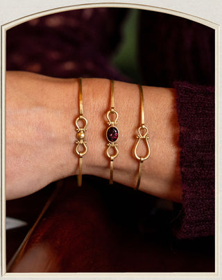 Three delicate gold bracelets displayed on a person's wrist. One bracelet features a small gold bead, another has an oval dark red gemstone, and the third is adorned with a gold infinity symbol.
