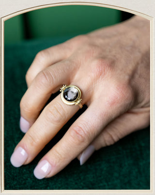 A gold ring with a dark oval gemstone worn on a person's ring finger, shown against a dark green textured background.