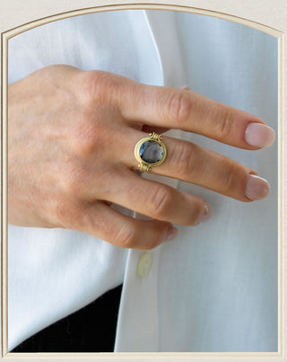 A close-up of a hand wearing a gold ring with a round dark gray stone, set against a white shirt.