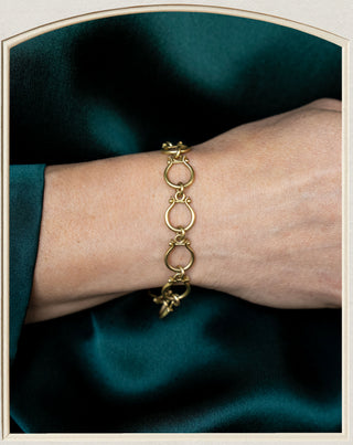 A gold-toned link bracelet with a small dangling charm, shown worn on a person's wrist against a dark teal satin fabric background.