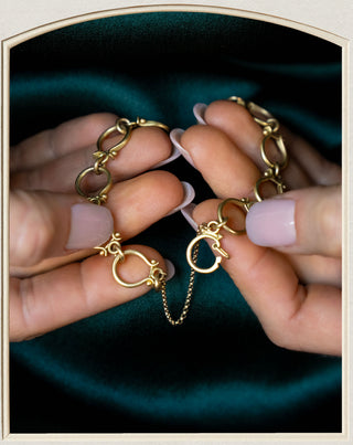 A close-up of a person's hands holding a gold chain bracelet with interlocking circular links and a short chain extension, set against a dark teal satin background.