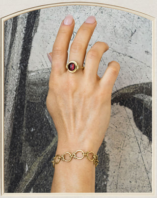A gold ring with a round dark red gemstone and a gold chain bracelet worn on a person's wrist, set against a textured gray background with a cream border.