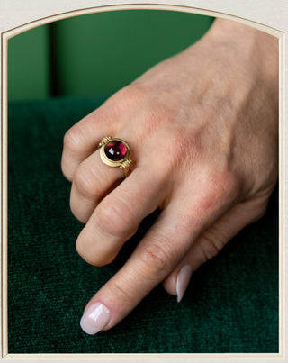 A gold signet ring with a round dark red gemstone, worn on a person's ring finger against a dark green textured background.