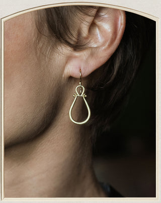 A close-up of a person's ear wearing a delicate gold teardrop-shaped dangle earring with a small decorative loop at the top.