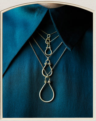 A layered silver necklace with multiple interlocking teardrop and oval pendants, displayed on a dark teal collared shirt against a light neutral background.