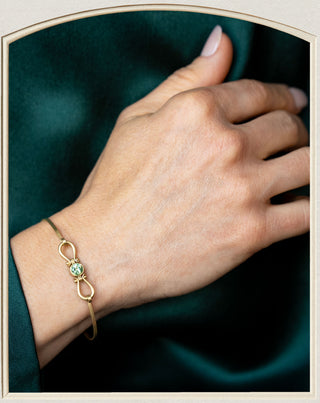 A gold infinity bracelet with a central green gemstone, worn on a person's wrist against a dark green satin background.
