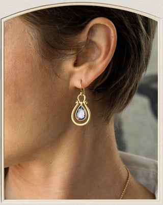 A close-up side view of a person's ear wearing a gold teardrop dangle earring with a pale blue teardrop gemstone.