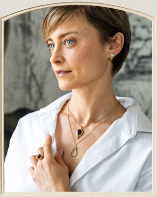 A woman wearing layered gold necklaces and a matching ring with dark oval gemstones, styled with a white collared shirt. The image is framed by a light beige arch border.