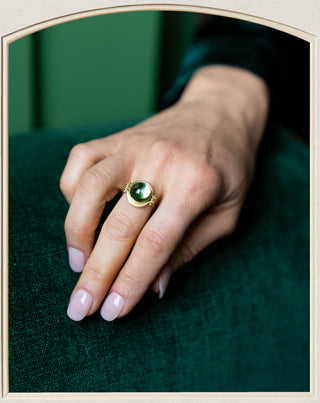 A gold ring with a round green cabochon gemstone worn on a person's finger, resting on a dark green textured surface.
