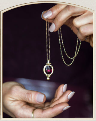 A gold pendant necklace with a round dark red gemstone is held between two hands with manicured nails against a dark background.